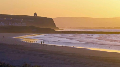 A view of the beach at Portstewart Strand at sunset, with walkers along the shoreline and a silhouette of the mainland in the background